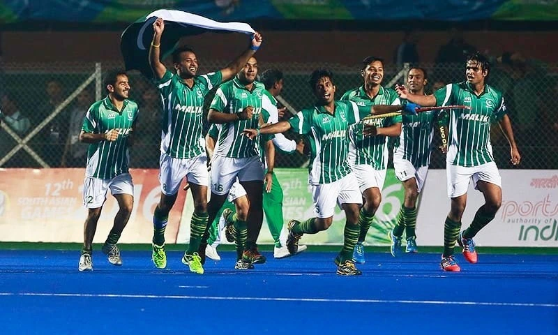 Pakistan hockey players in green jerseys celebrate on the field, holding the national flag.