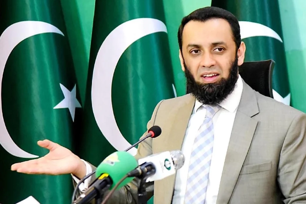 Information Minister Ataullah Tarar speaking at a press conference, seated in front of multiple Pakistan flags, with microphones placed on the desk