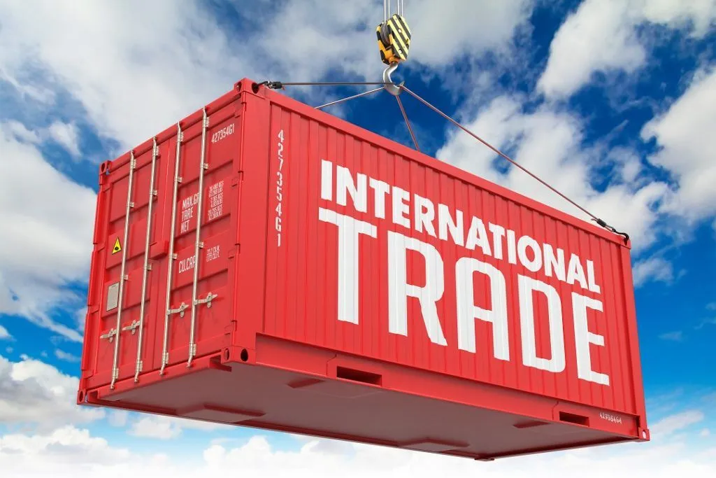 A large red shipping container labeled “INTERNATIONAL TRADE" being lifted by a crane against a bright blue sky with white clouds, symbolizing global commerce and cross-border trade.