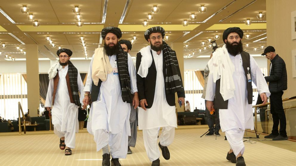 Group of Taliban officials walking in a large, well-lit conference hall, dressed in traditional Afghan attire with turbans and long robes, with media personnel and equipment visible in the background.