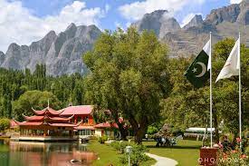 Scenic view of a Chinese-style building surrounded by lush greenery and a lake in Gilgit-Baltistan, with mountains in the background and Pakistani flags fluttering in the foreground.