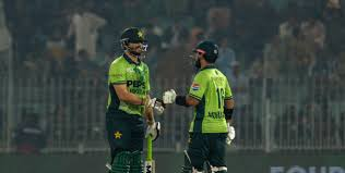 Two male Pakistani cricketers in green uniforms are fist-bumping on the field during a match.