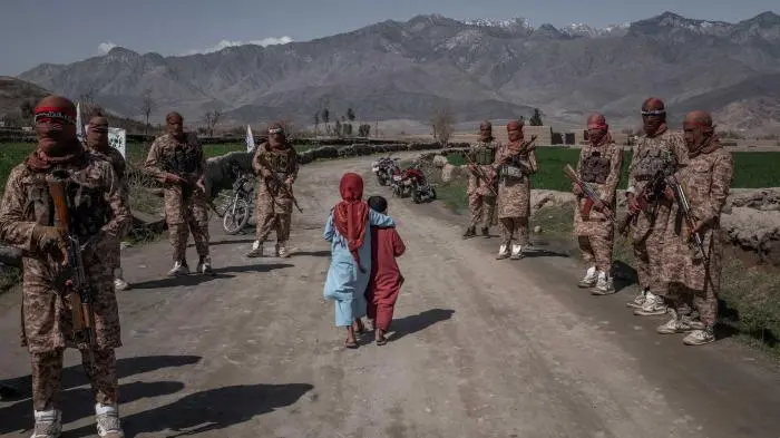 Two girls stand at the center surrounded by armed Afghan Taliban fighters, highlighting control and intimidation.